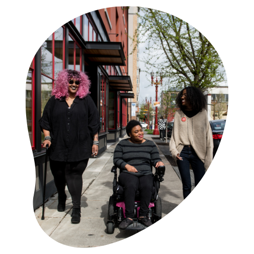 Three Black and disabled folx smile and stroll down a sidewalk side by side. On the left, a non-binary person walks with a cane in one hand and a tangle stim toy in the other. In the middle, a non-binary person rolls along in their power wheelchair. On the right, a femme gestures mid-walk.